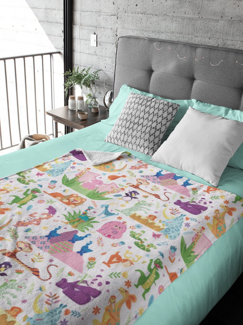May include: A white fleece blanket with a colorful pattern of cartoon animals and flowers. The blanket is on a bed with a light blue sheet and gray headboard.