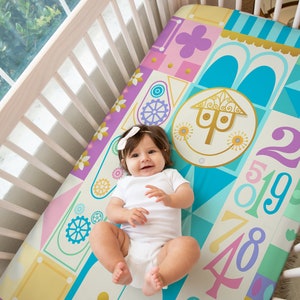 Op de afbeelding: Een baby-beddengoed met een kleurrijk geometrisch patroon met een fantasierijk huisontwerp, cijfers en tandwielen. Het beddengoed is in tinten blauw, roze, geel en groen.