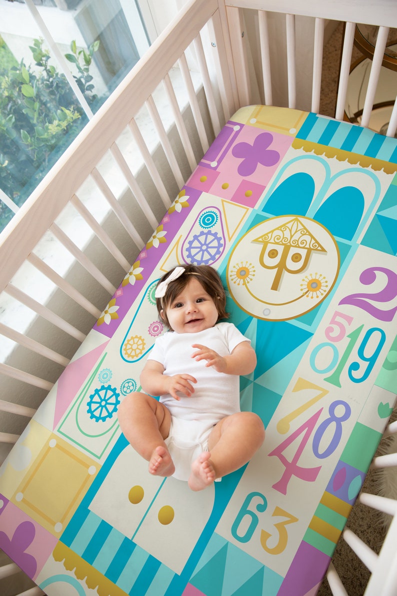 Op de afbeelding: Een baby-beddengoed met een kleurrijk geometrisch patroon met een fantasierijk huisontwerp, cijfers en tandwielen. Het beddengoed is in tinten blauw, roze, geel en groen.
