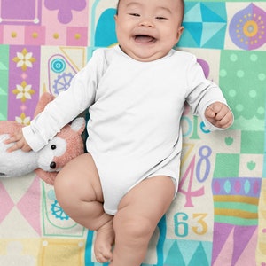Op de afbeelding: Een lachende baby in een wit, lang mouwig onesie ligt op een kleurrijk, gepatroneerd deken. Het deken heeft verschillende vormen, kleuren en nummers.
