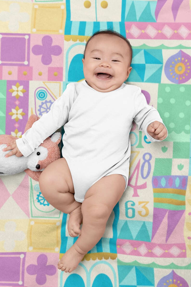Op de afbeelding: Een lachende baby in een wit, lang mouwig onesie ligt op een kleurrijk, gepatroneerd deken. Het deken heeft verschillende vormen, kleuren en nummers.