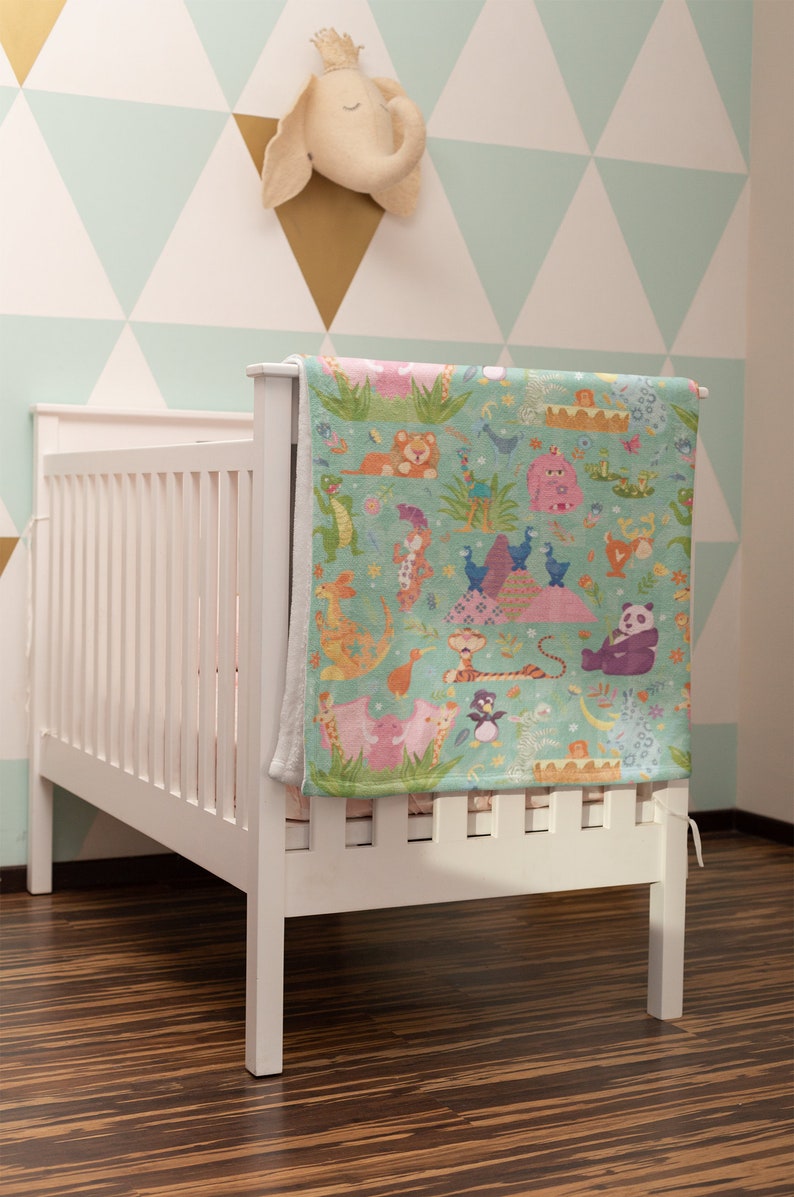 May include: A white crib with a colorful baby blanket featuring a variety of animals, including a lion, a penguin, a panda, and a giraffe. The blanket is hanging on the crib and has a light blue background with green grass and flowers.