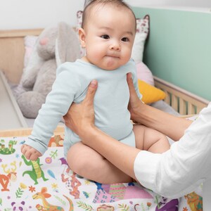 May include: A baby wearing a light blue onesie is being held by a person. The baby is sitting on a white blanket with a colorful animal print. The blanket features a variety of animals, including a panda, a tiger, a kangaroo, and a penguin.