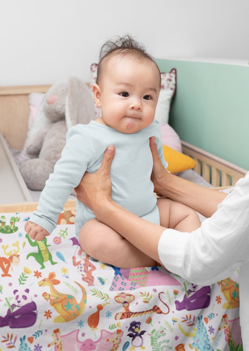 May include: A baby wearing a light blue onesie is being held by a person. The baby is sitting on a white blanket with a colorful animal print. The blanket features a variety of animals, including a panda, a tiger, a kangaroo, and a penguin.
