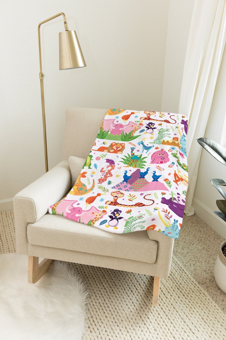 May include: A white throw blanket with a colorful pattern of animals and flowers. The animals include a lion, a tiger, a penguin, a rhinoceros, a giraffe, and an elephant. The blanket is on a beige armchair.