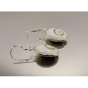 May include: Silver earrings with a white, circular design and a central spiral pattern. The earrings have a hook fastening and are shown on a reflective surface. The earrings are a piece of jewellery.