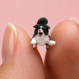 Puede incluir: Una figura en miniatura de un perro blanco y negro con la cola enroscada y la boca abierta mostrando una lengua rosa. La pequeña figura canina se muestra en una postura juguetona, inclinada hacia adelante con las patas delanteras extendidas.
