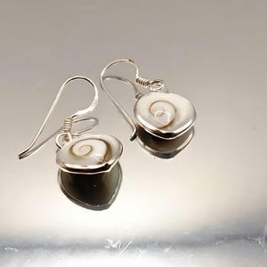 May include: A pair of drop earrings featuring polished white and brown spiral shell centers set in silver-toned metal frames. Each earring hangs from a curved silver-toned wire hook, showcasing a natural swirl pattern on the circular shell surface.