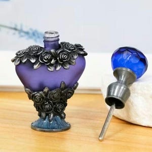 May include: A heart-shaped purple glass perfume bottle featuring a metallic silver rose motif around the neck and base. The bottle includes a separate stopper with a faceted blue glass orb top and a long metal applicator wand attached to the base.