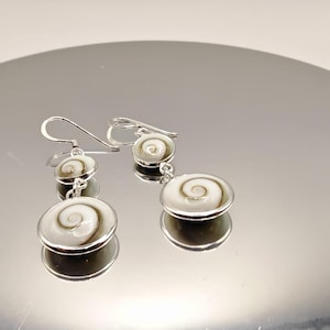 May include: Silver earrings with a spiral design. Each earring has two circular pendants, one small and one large, with a white and tan spiral pattern. The earrings hang from silver hooks.