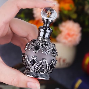 May include: A small, round perfume bottle with a frosted glass body encased in an ornate, dark metallic filigree overlay featuring black gemstone accents. The bottle is topped with a clear, faceted crystal stopper and a dark metallic neck.