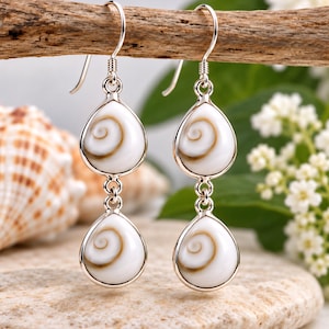 May include: A pair of drop earrings featuring two teardrop-shaped Shiva shell cabochons set in polished silver. Each earring consists of two stacked shells with a natural spiral pattern in white and brown, suspended from a silver hook wire.