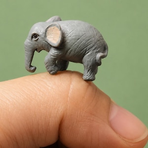 May include: A miniature grey elephant figurine with a textured surface, visible skin folds, and light tan inner ears. The small sculpture features a curved trunk, a rounded body, and a short tail, showcasing detailed craftsmanship in a compact form.