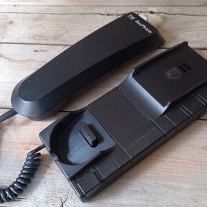 Puede incluir: Un teléfono negro con cable con una unidad base y un auricular. La unidad base tiene una superficie texturizada y el auricular tiene el texto "T&M BusPhone" en la parte superior.