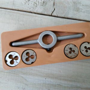 Può includere: Un set di maschi e filiere in metallo, con un manico argentato e diverse filiere circolari. Il set è alloggiato in una custodia rettangolare di plastica arancione. Le filiere hanno un design a tre lobi.