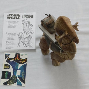 Op de afbeelding: Een bruin plastic Star Wars Ronto speelgoed met een zadel en een Jawa figuur. Het speelgoed ligt op een wit oppervlak met instructies en een vel stickers.