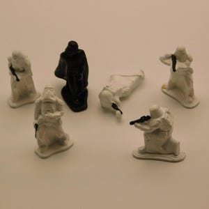 May include: A collection of six small plastic figurines including five white stormtrooper soldiers and one black Darth Vader figure. The figures are depicted in various poses, with some standing, one kneeling, and one lying down on the ground.