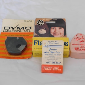 May include: A collection of vintage office supplies, including black DYMO embossing tape, a GE promotional card with a child and teddy bear, a box of Flas bulbs, a Garrett and MacInnis Personnel Agency first aid kit, and a pink Touch and Go typing correction tape.