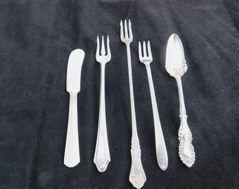 May include: A collection of silver-colored flatware, including a butter knife, three forks, and a serving spoon. The utensils are arranged on a dark background. The spoon and one fork have ornate handle designs.