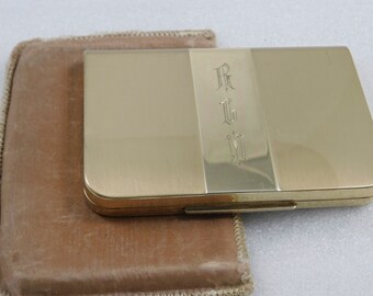 May include: A gold-colored metal compact with a velvet pouch. The compact has a vertical band with the initials "R.G.M." engraved on it.