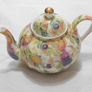 May include: A white teapot with a gold lid and a fruit pattern. The pattern includes lemons, grapes, and other fruits in shades of yellow, purple, and pink.