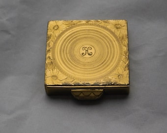 May include: A gold-colored compact with a floral design and a monogrammed initial "K" in the center.