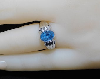 May include: A silver ring with a blue oval-shaped gemstone set in the centre. The ring has a decorative design on the band.