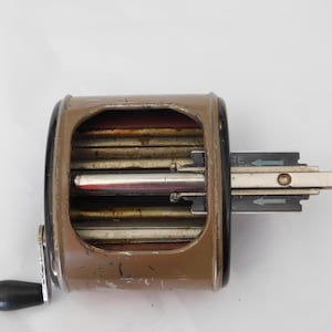 Puede incluir: Un sacapuntas vintage marrón y negro con manivela y mecanismo deslizante. El sacapuntas tiene forma rectangular con una serie de cuchillas visibles en el interior. La palabra "RATE" está impresa en el mecanismo deslizante.