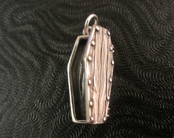May include: A silver-colored coffin pendant with a detailed wood grain design. The coffin is open and has a black interior.