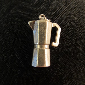 May include: A small, silver-colored charm in the shape of a stovetop espresso maker, also known as a Moka pot. The charm has a handle and a loop at the top for attaching to a chain or bracelet. The background is a textured black surface.