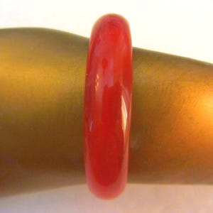 May include: A vibrant red bangle bracelet. The smooth, glossy surface reflects light, highlighting its rounded shape. The bracelet is shown on a golden-toned surface, creating a striking contrast.