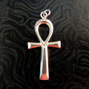 May include: A polished silver-toned ankh pendant featuring a classic looped top and a vertical cross bar. The metal surface is smooth and reflective, showcasing a clean, metallic finish with a small circular jump ring attached at the very top.