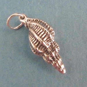 May include: A detailed silver seashell charm with a textured surface and a small loop for attachment. The charm is elongated and tapered, with intricate ridges and a polished finish. The background is a solid light blue.