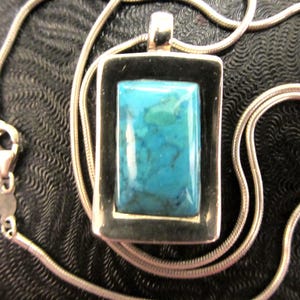 May include: A silver necklace featuring a rectangular pendant with a vibrant turquoise stone. The stone is set within a dark frame, and the necklace has a delicate, snake-like chain. The clasp is visible.