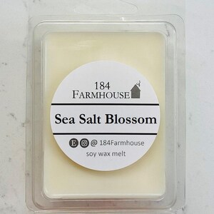 May include: A rectangular soy wax melt in a clear plastic clamshell. The wax is a pale yellow color. A white circular label reads "184 Farmhouse Sea Salt Blossom" with social media handles.