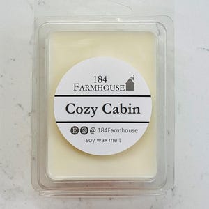 May include: A rectangular soy wax melt in a clear plastic clamshell. The wax is a pale yellow color. A white circular label reads "184 Farmhouse Cozy Cabin" with social media handles. The product is designed for home fragrance.
