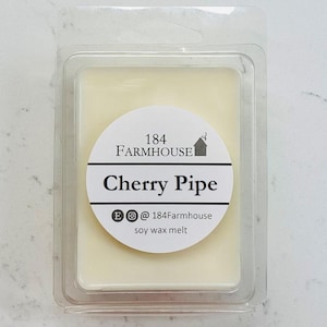 May include: A clear plastic container holding a light yellow soy wax melt. The wax melt has a circular label with the text "184 Farmhouse Cherry Pipe" and social media handles. The product is on a white surface.