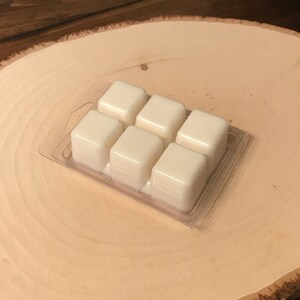 May include: A clear plastic clamshell package containing six ivory-colored wax melt cubes. The cubes are arranged in two rows of three. The package sits on a light-colored, wooden, circular surface with a natural wood grain.