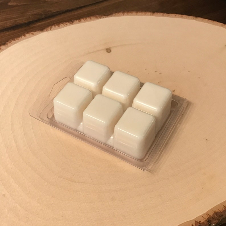 May include: A clear plastic clamshell package containing six ivory-colored wax melt cubes. The cubes are arranged in two rows of three. The package sits on a light-colored, wooden, circular surface with a natural wood grain.