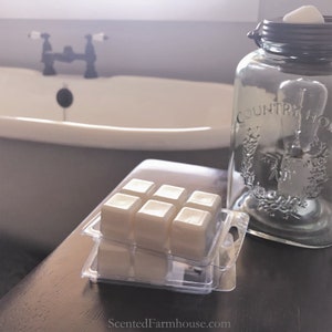 May include: A clear glass jar with a metal lid and the words "COUNTRY HOME" printed on the side. Several wax melt cubes are in a clear plastic container. The background includes a white bathtub and a faucet.