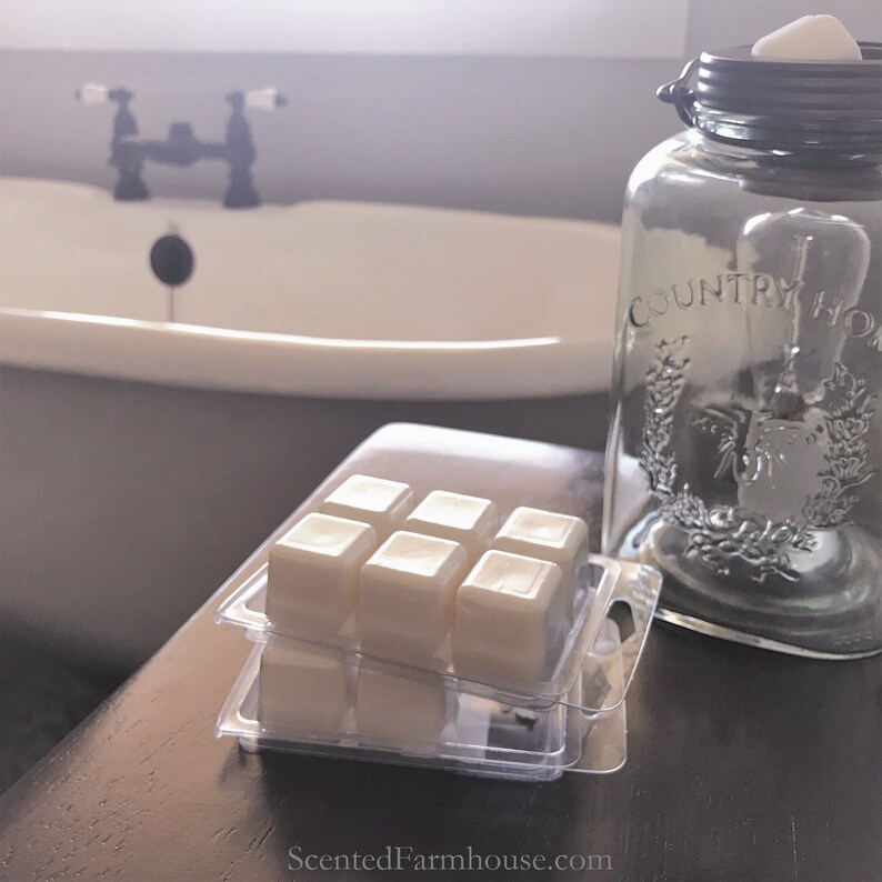 May include: A clear glass jar with a metal lid and the words "COUNTRY HOME" printed on the side. Several wax melt cubes are in a clear plastic container. The background includes a white bathtub and a faucet.