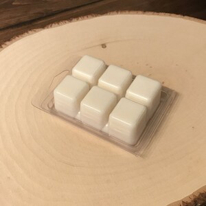 May include: A clear plastic clamshell container holding six ivory-colored wax melt cubes. The wax melts are arranged in two rows of three, with a glossy, smooth surface. The container sits on a light brown, wooden, circular surface.