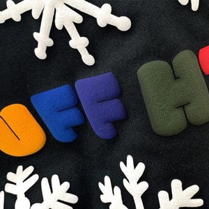 May include: A black sweatshirt with the words "PUFF HTV" in colorful, puffy letters. The letters are in different colors, including brown, orange, blue, purple, green, and red. The sweatshirt is decorated with white snowflake designs.