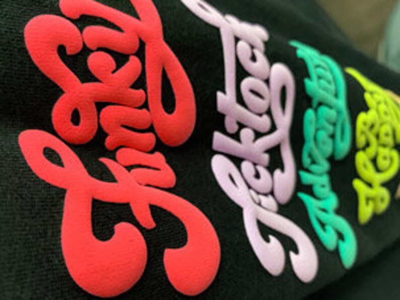 May include: A black t-shirt with five different colored words printed on it. The words are "Funky", "Ticktock", "Adventure", "Free", and "Love".