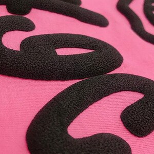 May include: Close-up of a pink fabric with black raised lettering that reads "Coco".