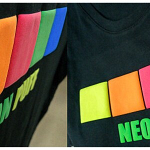 May include: Black t-shirt with a neon puff design. The design features a row of five colorful squares in yellow, orange, red, green, and blue. The text "NEON PUFF" is printed in yellow below the squares.