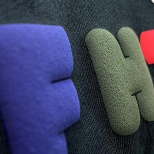 May include: Close-up of a black t-shirt with the word "BE" in blue, "HT" in green, and "V" in red. The letters are raised and have a textured surface.