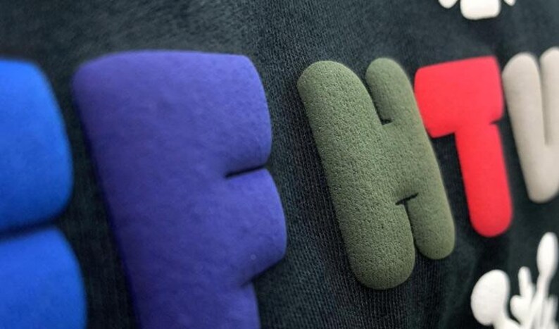 May include: Close-up of a black t-shirt with the word "BE" in blue, "HT" in green, and "V" in red. The letters are raised and have a textured surface.