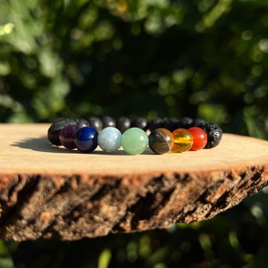 May include: A beaded bracelet featuring seven chakra stones in a rainbow of colors, including purple, blue, light blue, green, yellow, orange, and red. The bracelet also includes black lava beads. The bracelet is displayed on a wooden surface.