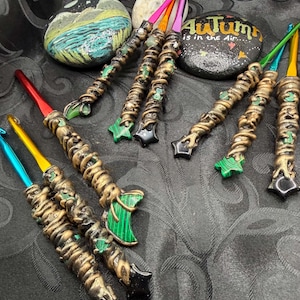 May include: A collection of metal crochet hooks with custom sculpted handles featuring metallic bronze, gold, and black textures. The handles are adorned with green malachite-style crescent moons and star shapes. Two painted rocks are visible, one with a landscape and one with the text Autumn is in the Air.